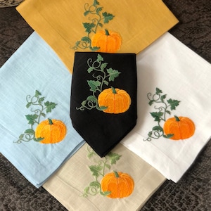 May include: Four embroidered linen napkins featuring a pumpkin and vine design. The napkins are in the colours blue, white, beige, and black.