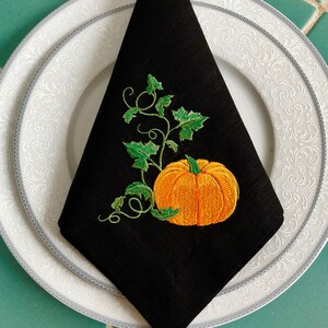 May include: Black linen napkin featuring an embroidered pumpkin and vine design. The pumpkin is orange with a green stem and leaves. The vine is green with leaves.
