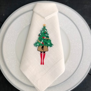 May include: White linen napkin featuring an embroidered design of a person holding a Christmas tree. The tree is green with red baubles and a gold star on top. The person is wearing a brown coat and red boots.