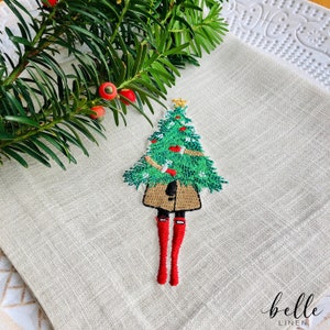 May include: A beige linen kitchen towel with an embroidered design of a person wearing a brown coat and red boots holding a green Christmas tree with red baubles. The text "belle linen" is embroidered in the bottom right corner.