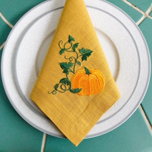 K&ouml;nnte beinhalten: Eine gefaltete gelbe Serviette mit einer gestickten orangefarbenen K&uuml;rbis- und gr&uuml;nen Ranken-Design, platziert auf einem wei&szlig;en Teller mit silbernem Rand. Die Serviette besteht aus einem strukturierten Stoff, und der Teller hat ein zartes Blumenmuster.
