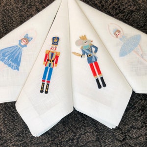 Puede incluir: Cuatro servilletas de lino blanco con imágenes bordadas de una bailarina, un cascanueces, un rey ratón y una segunda bailarina. La bailarina lleva un vestido azul y blanco y el cascanueces lleva un uniforme rojo y azul.