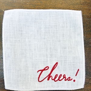 May include: White linen handkerchief with the word "Cheers!" embroidered in red script. The handkerchief has a textured weave and a simple, elegant design, perfect for a gift or special occasion.
