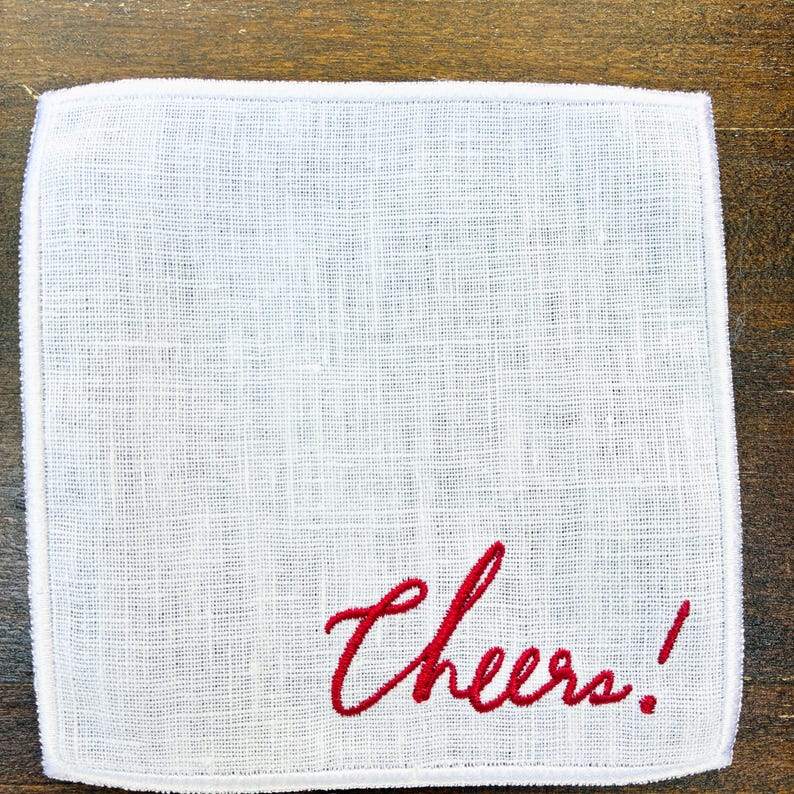May include: White linen handkerchief with the word "Cheers!" embroidered in red script. The handkerchief has a textured weave and a simple, elegant design, perfect for a gift or special occasion.
