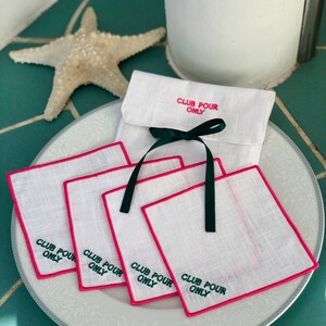 May include: Set of four white linen coasters with pink edging and green embroidery that reads "Club Pour Only". The coasters are stacked on a white plate with a decorative pattern. The coasters are packaged in a white linen pouch with a green ribbon.