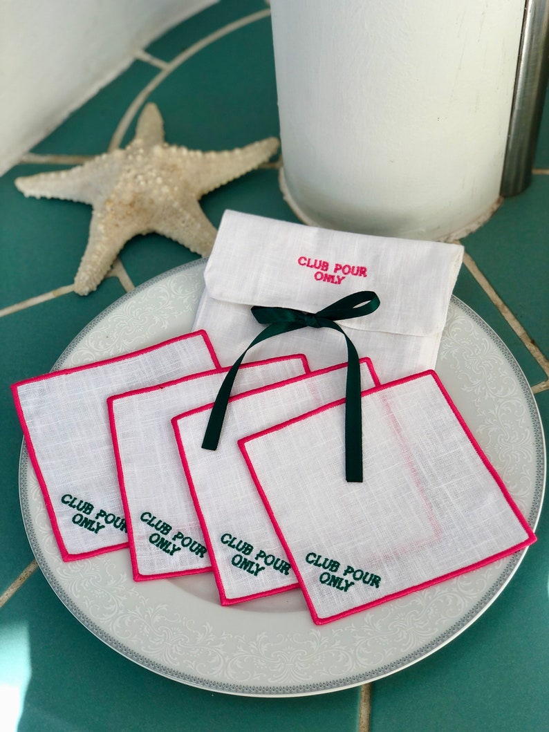 May include: Set of four white linen coasters with pink edging and green embroidery that reads "Club Pour Only". The coasters are stacked on a white plate with a decorative pattern. The coasters are packaged in a white linen pouch with a green ribbon.