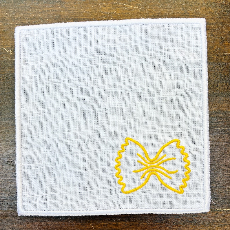 May include: White linen square with a yellow embroidered bow tie pasta design. The square has a white border and is set against a dark wood surface. The pasta design is in the lower right corner.