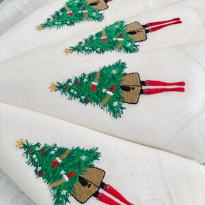 May include: Four white linen napkins with embroidered Christmas trees and figures wearing red stockings. Each napkin features a green Christmas tree with brown ornaments and a figure with red stockings and a brown skirt.