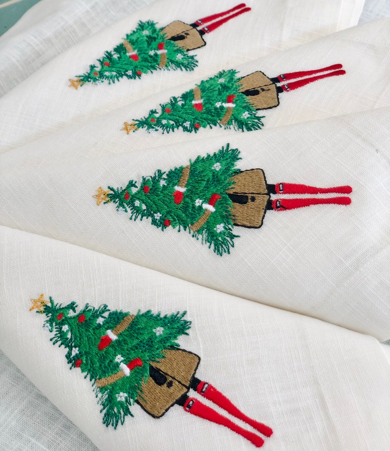 May include: Four white linen napkins with embroidered Christmas trees and figures wearing red stockings. Each napkin features a green Christmas tree with brown ornaments and a figure with red stockings and a brown skirt.