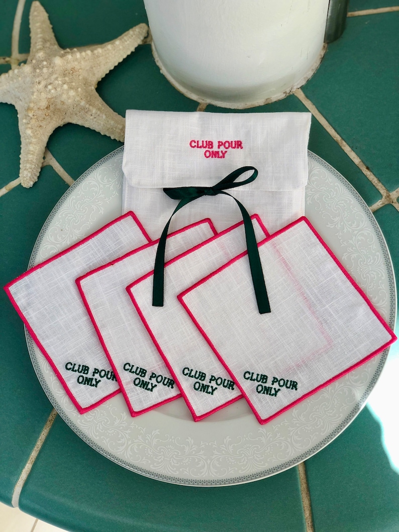 May include: Set of four white linen napkins with pink edging and green embroidery that reads "Club Pour Only". The napkins are folded and stacked on a white plate with a white linen pouch with the same embroidery on top.