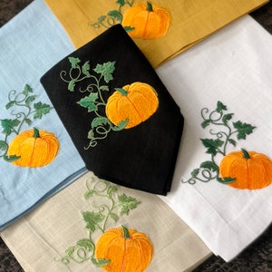 May include: Set of four embroidered linen napkins featuring a pumpkin vine design. The napkins are in various colours, including blue, white, black, and beige.