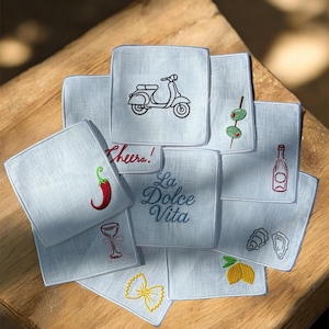 May include: A collection of light blue square embroidered cocktail napkins. Designs include a scooter, olives, a bottle, a chili pepper, a wine glass, pasta, a lemon, and the phrase "La Dolce Vita".