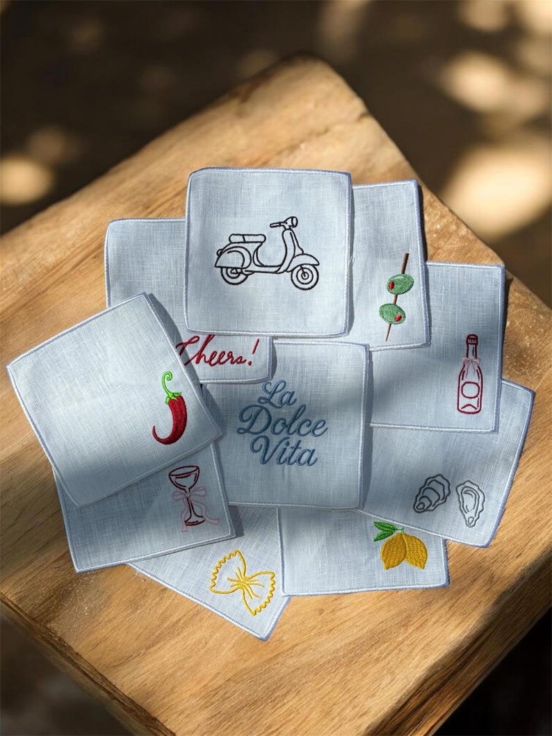 May include: A collection of light blue square embroidered cocktail napkins. Designs include a scooter, olives, a bottle, a chili pepper, a wine glass, pasta, a lemon, and the phrase "La Dolce Vita".