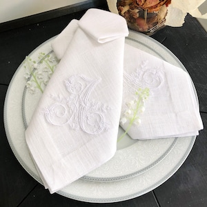 Op de afbeelding: Twee witte linnen servetten met geborduurde bloemenmotieven en de letter "A" in een schrijfletter. De servetten zijn gevouwen en op een wit bord met een zilveren rand geplaatst.