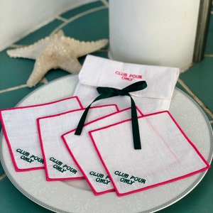 May include: Set of five white linen coasters with pink edging and green embroidery that reads "Club Pour Only". The coasters are arranged on a white plate with a decorative border.