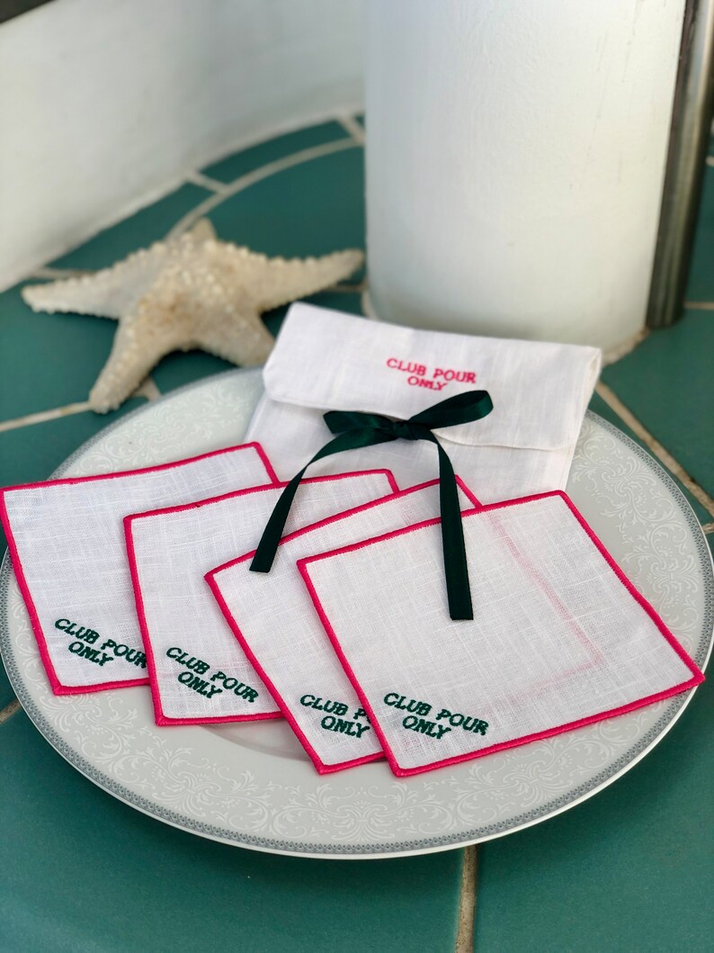 May include: Set of five white linen coasters with pink edging and green embroidery that reads "Club Pour Only". The coasters are arranged on a white plate with a decorative border.