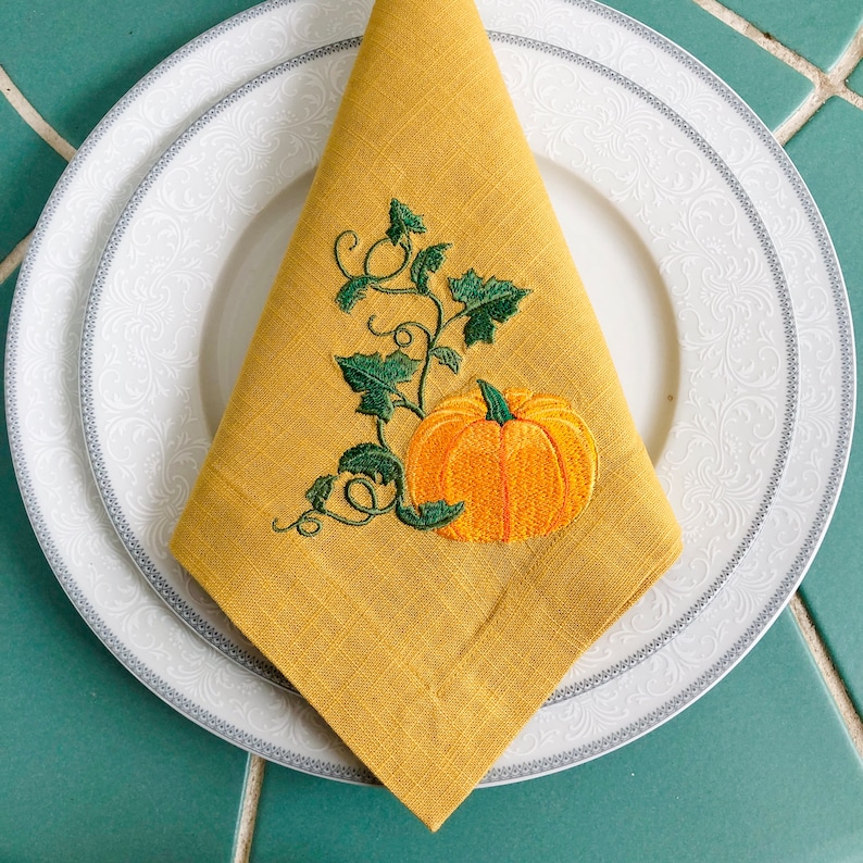 May include: A yellow linen napkin with an embroidered pumpkin and vine design. The pumpkin is orange with a green stem and leaves.