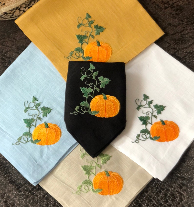 May include: Four embroidered linen napkins featuring a pumpkin and vine design. The napkins are in the colours blue, white, beige, and black.