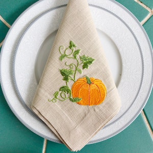 May include: A beige linen napkin with an embroidered design of a green pumpkin vine and an orange pumpkin.