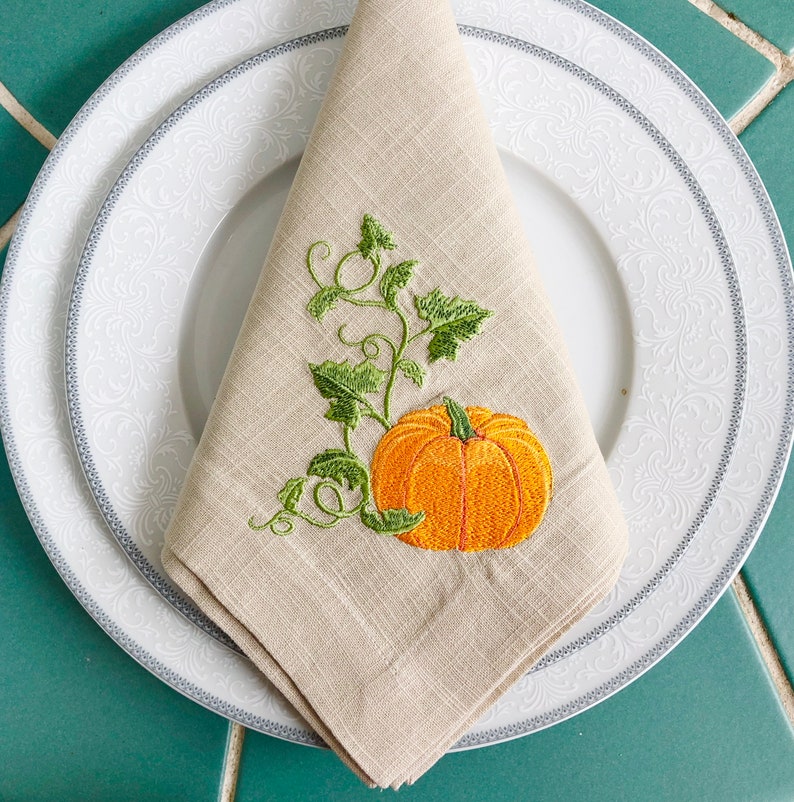 May include: A beige linen napkin with an embroidered design of a green pumpkin vine and an orange pumpkin.