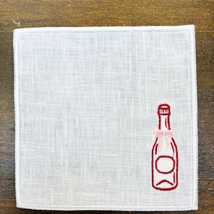 May include: White linen napkin with a red embroidered champagne bottle design and a pink bow. The napkin has a simple, elegant design, perfect for a special occasion or everyday use. The napkin is square-shaped.