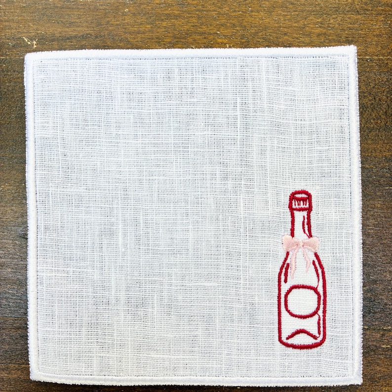 May include: White linen napkin with a red embroidered champagne bottle design and a pink bow. The napkin has a simple, elegant design, perfect for a special occasion or everyday use. The napkin is square-shaped.