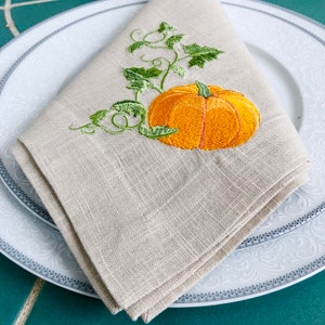 May include: A beige linen napkin with an embroidered orange pumpkin and green leaves.
