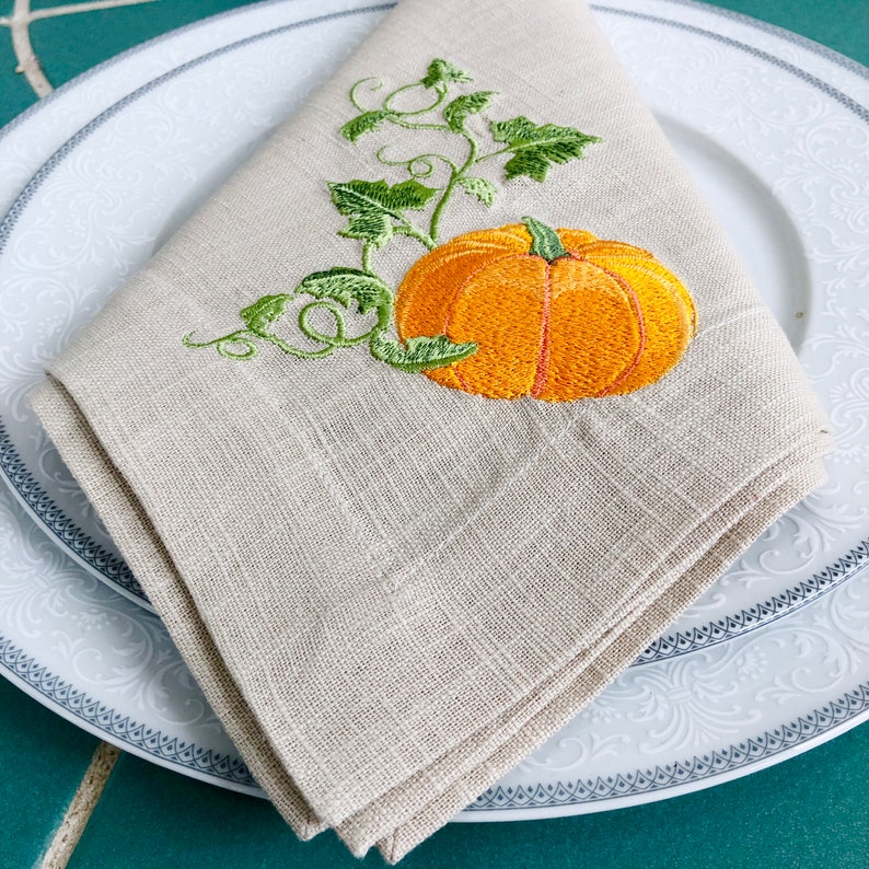May include: A beige linen napkin with an embroidered orange pumpkin and green leaves.