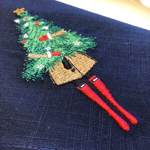 May include: Embroidered design on a dark blue fabric featuring a person holding a Christmas tree with red baubles and a star on top. The person is wearing a brown coat and red boots.