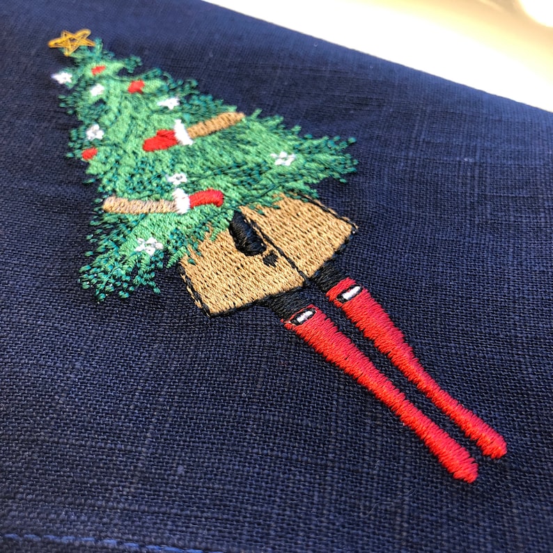 May include: Embroidered design on a dark blue fabric featuring a person holding a Christmas tree with red baubles and a star on top. The person is wearing a brown coat and red boots.