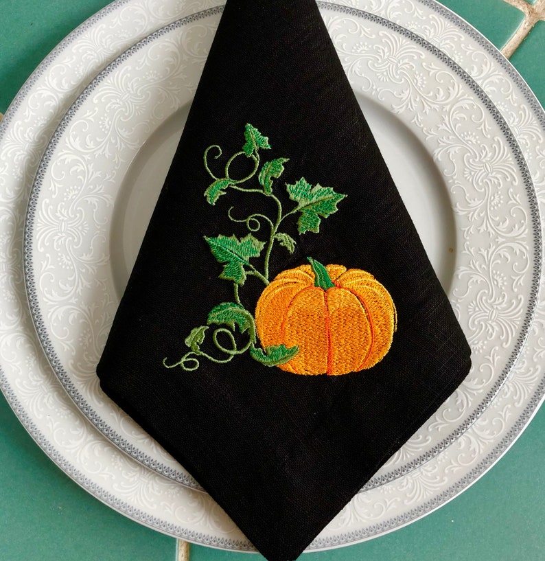May include: Black linen napkin featuring an embroidered pumpkin and vine design. The pumpkin is orange with a green stem and leaves. The vine is green with leaves.