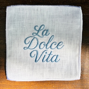 May include: White linen handkerchief with the embroidered phrase "La Dolce Vita" in a script font. The text is in a light blue color. The handkerchief has a square shape and a delicate, textured appearance.