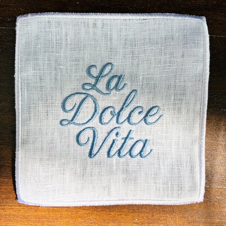 May include: White linen handkerchief with the embroidered phrase "La Dolce Vita" in a script font. The text is in a light blue color. The handkerchief has a square shape and a delicate, textured appearance.
