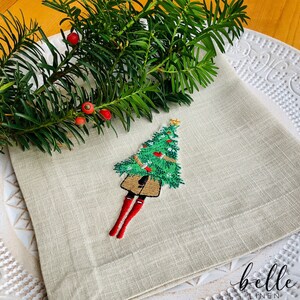 May include: A beige linen napkin featuring an embroidered design of a Christmas tree wearing red boots. The tree is decorated with green and red baubles. The napkin is folded in thirds and has a "belle linen" logo in the bottom right corner.