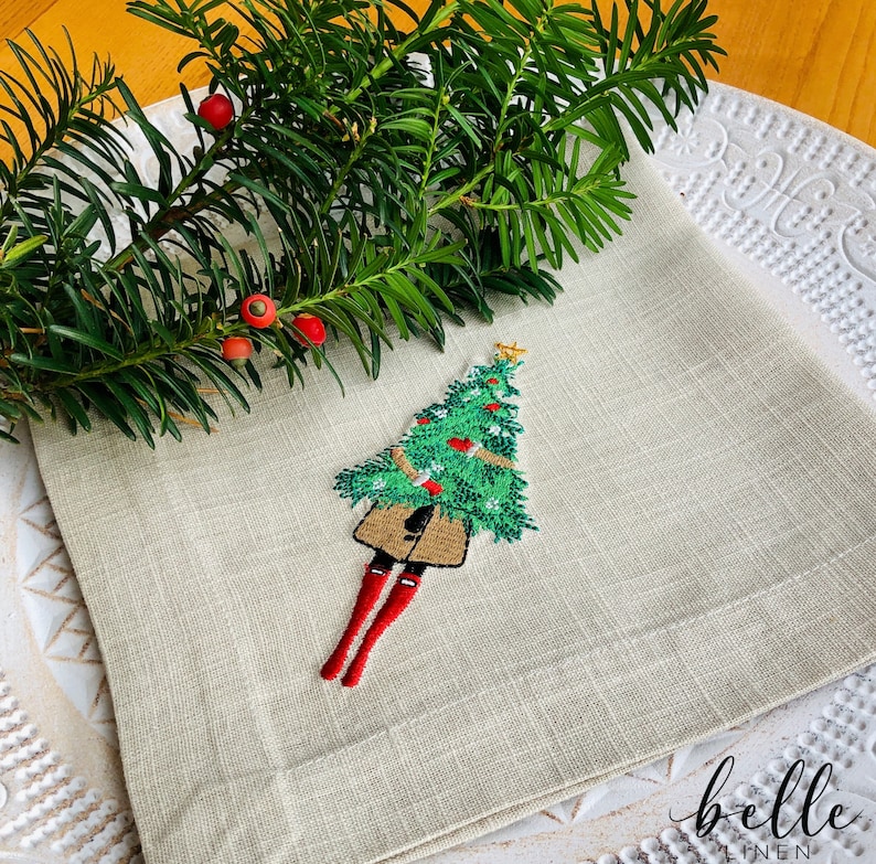 May include: A beige linen napkin featuring an embroidered design of a Christmas tree wearing red boots. The tree is decorated with green and red baubles. The napkin is folded in thirds and has a "belle linen" logo in the bottom right corner.