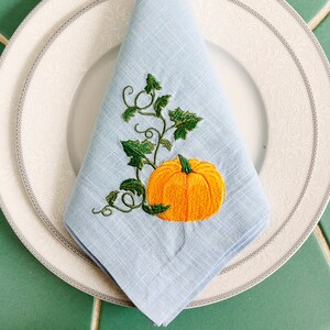 May include: A light blue linen napkin with an embroidered pumpkin and vine design. The pumpkin is orange with a green stem and leaves.