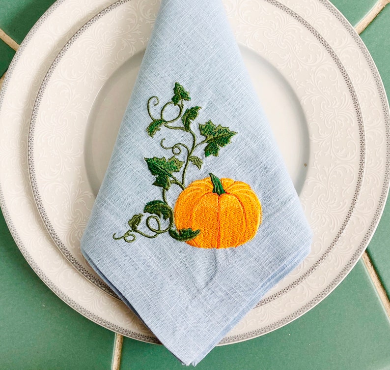 May include: A light blue linen napkin with an embroidered pumpkin and vine design. The pumpkin is orange with a green stem and leaves.
