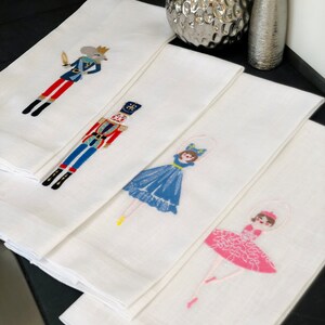 May include: Set of four white kitchen towels with embroidered images of a nutcracker, a ballerina, and a prince. The towels are folded and stacked on top of each other.