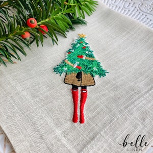 May include: Embroidered linen kitchen towel featuring a girl holding a Christmas tree. The girl is wearing a brown coat and red boots. The towel has the text "belle linen" in the bottom right corner.
