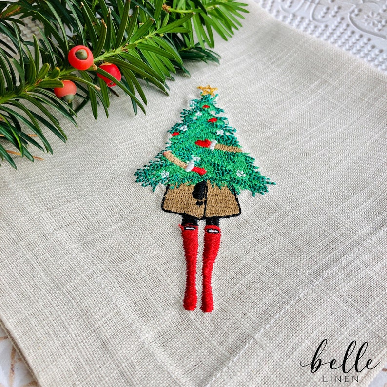 May include: Embroidered linen kitchen towel featuring a girl holding a Christmas tree. The girl is wearing a brown coat and red boots. The towel has the text "belle linen" in the bottom right corner.