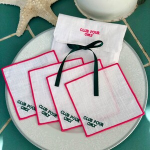 May include: Four white linen napkins with pink edging and green embroidery that reads "Club Pour Only". The napkins are stacked on a white plate with a decorative pattern. A white linen pouch with green ribbon and the same embroidery is in the background.