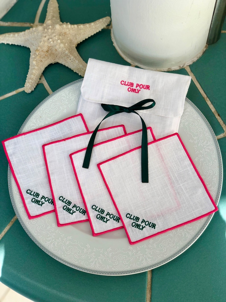 May include: Four white linen napkins with pink edging and green embroidery that reads "Club Pour Only". The napkins are stacked on a white plate with a decorative pattern. A white linen pouch with green ribbon and the same embroidery is in the background.