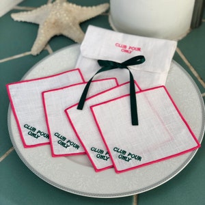 May include: Four white linen cocktail napkins with pink edging and green embroidery that reads "Club Pour Only". The napkins are stacked on a white plate with a decorative pattern.
