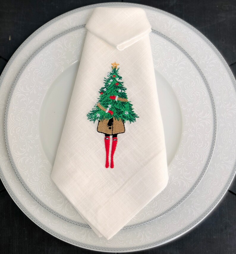 May include: White linen napkin featuring an embroidered design of a person holding a Christmas tree. The tree is green with red baubles and a gold star on top. The person is wearing a brown coat and red boots.