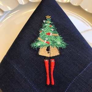 May include: A navy blue linen napkin featuring an embroidered design of a person wearing a brown coat and red boots holding a green Christmas tree.