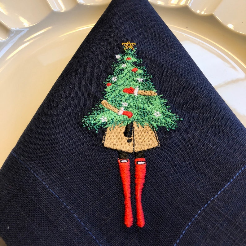 May include: A navy blue linen napkin featuring an embroidered design of a person wearing a brown coat and red boots holding a green Christmas tree.