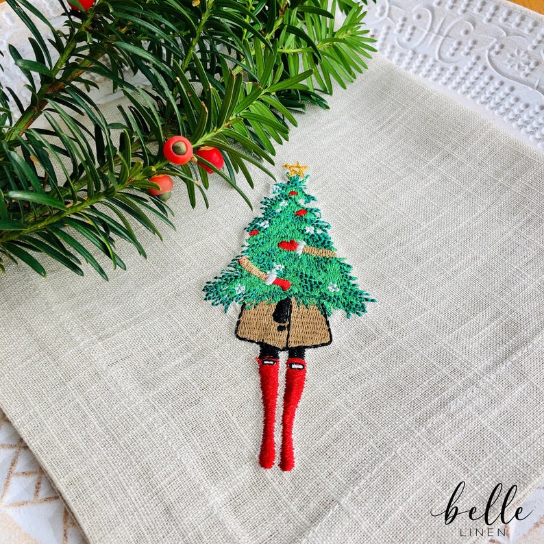 May include: A beige linen kitchen towel with an embroidered design of a person wearing a brown coat and red boots holding a green Christmas tree with red baubles. The text "belle linen" is embroidered in the bottom right corner.