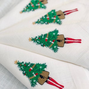 May include: Set of four white linen napkins featuring embroidered Christmas trees and figures with red legs. Each napkin has a green Christmas tree with brown decorations and a gold star on top.