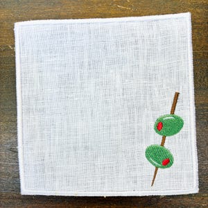 May include: White linen cocktail napkin with an embroidered design of two green olives with red centers on a wooden skewer. The napkin has a white stitched border and is set against a wooden background.
