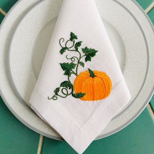 May include: White linen napkin featuring an embroidered pumpkin and vine design. The pumpkin is orange with a green stem and leaves. The vine is green with leaves.